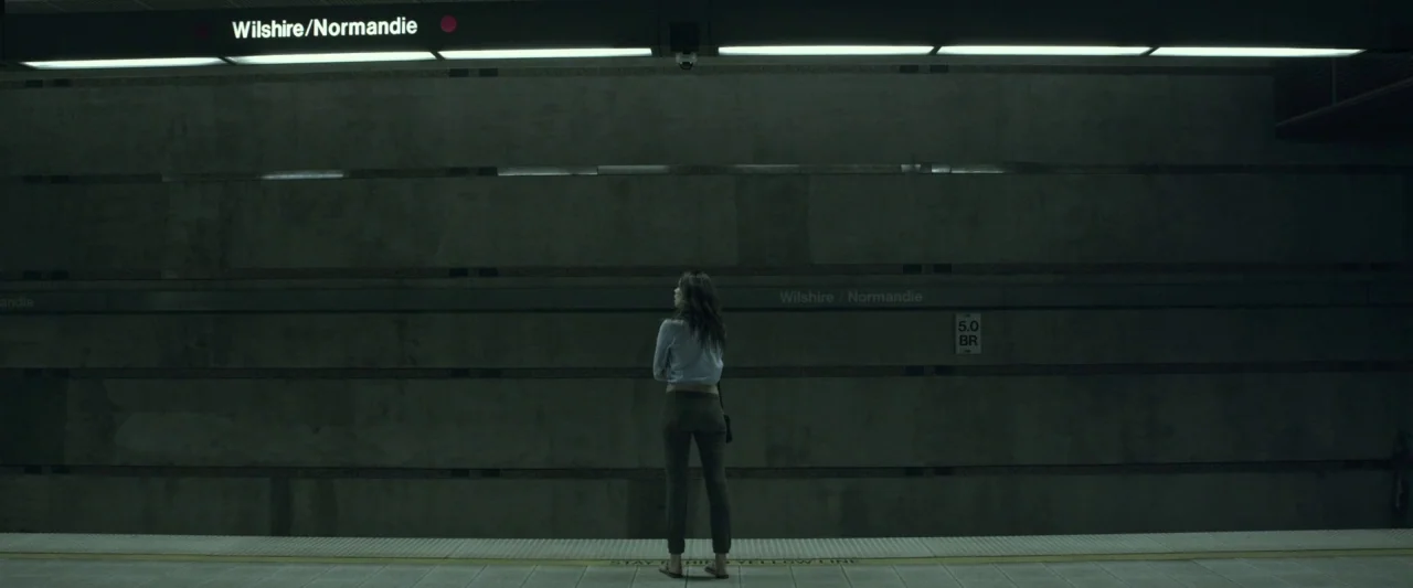 Starry Eyes (2014) movie still: wide — A stark subway platform features gray concrete walls and a prominent overhead transit sign. A single woman…