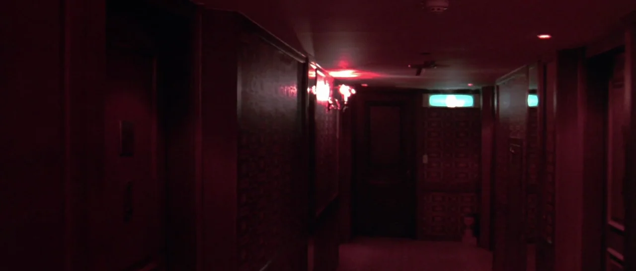 Oldboy (2003) movie still: establishing — A dim, narrow hotel hallway features patterned wallpaper and several closed wooden doors. A faint teal…