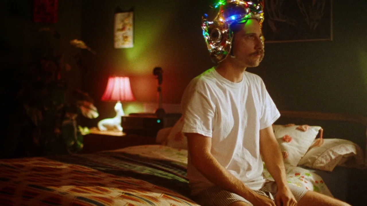 Strawberry Mansion (2021) movie still: medium — A dimly lit room with olive green walls contains a bed covered in patterned textiles. A man with a…