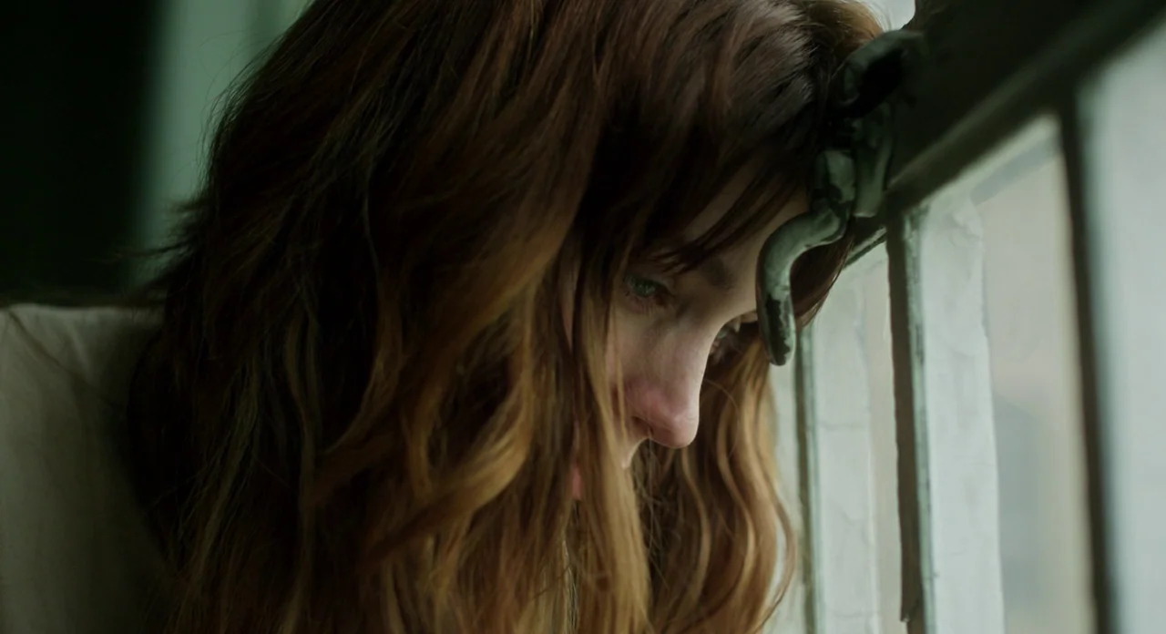 Side Effects (2013) movie still: closeup — A woman leans against the metal latch of a white-framed window. Soft daylight illuminates her face as she…