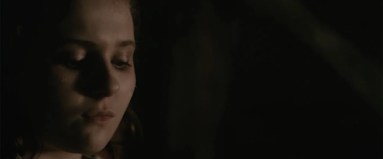 Maggie (2015) movie still: closeup — A dimly lit room with heavy shadows surrounding the subject. A young woman with brown hair keeps her eyes…