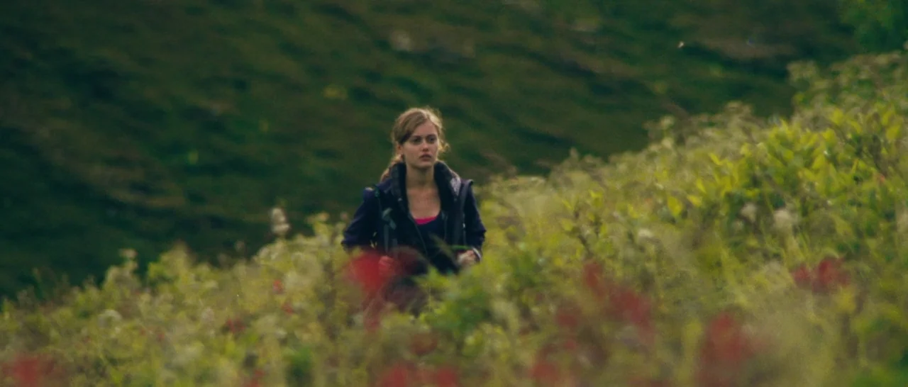 Wildlike (2014) movie still: medium — A woman walks through a grassy hillside covered in low-lying green and red shrubs. The rolling slope stretches…