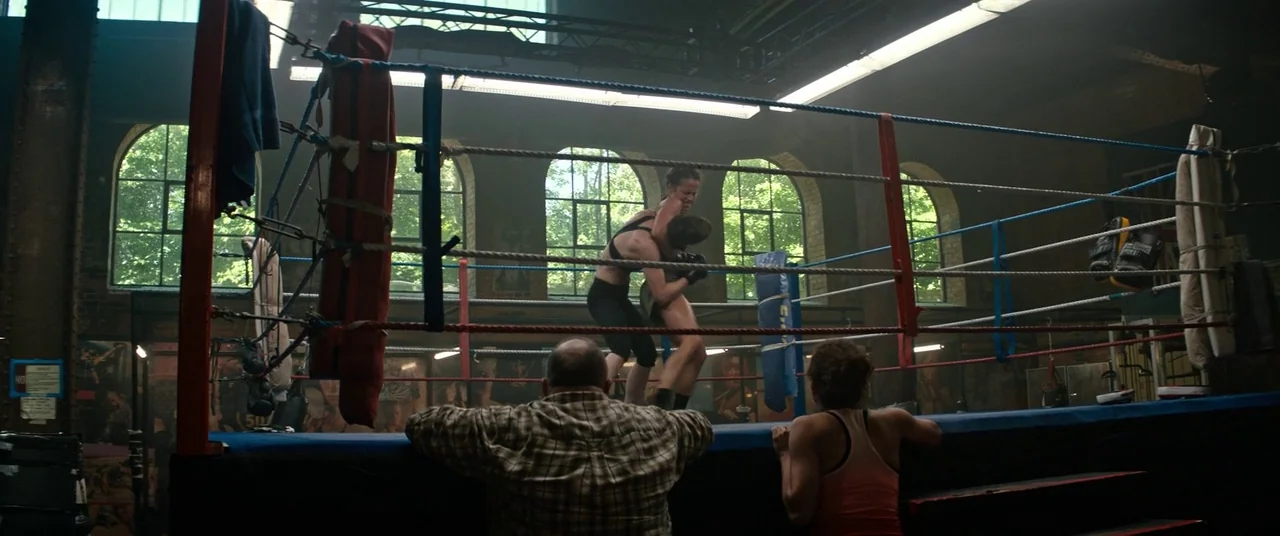 Tomb Raider (2018) movie still: wide — An indoor gym featuring a central elevated fighting ring surrounded by brick walls. Two women practice…