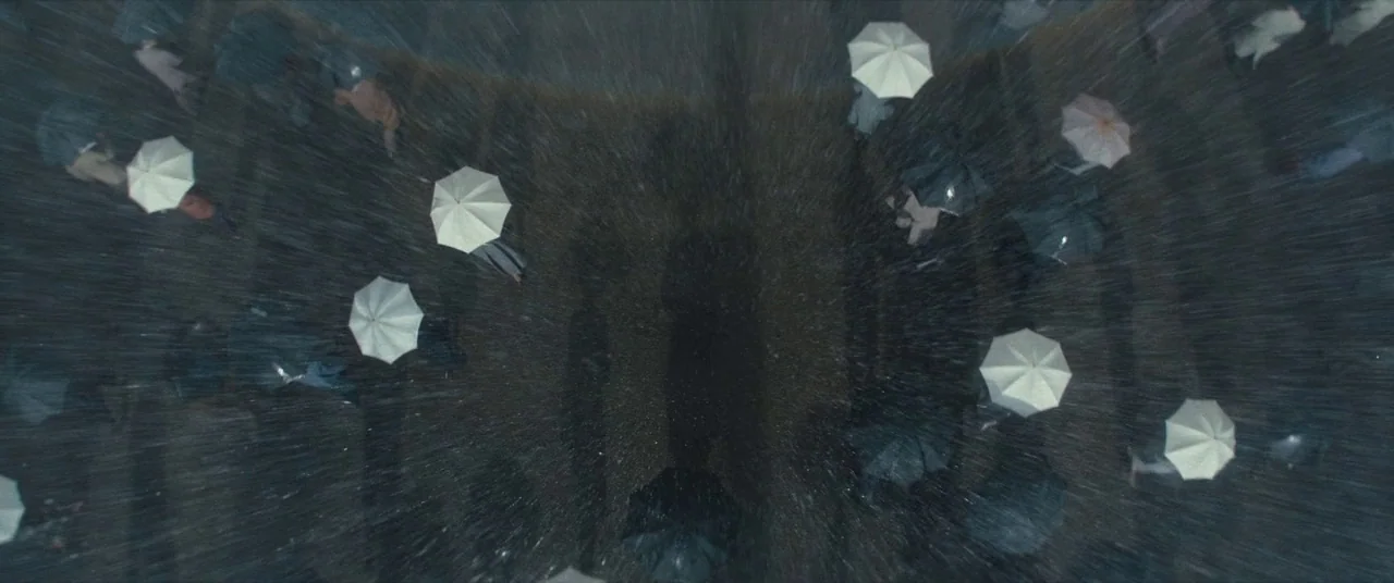 Christopher Robin (2018) movie still: establishing — A top-down view shows a crowd of people navigating a dark street during rain. Numerous…