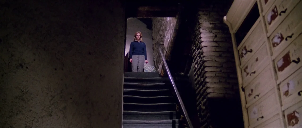 The New York Ripper (1982) movie still: wide — A dark, narrow staircase with exposed stone walls leads up to a bright doorway. A woman stands in the…