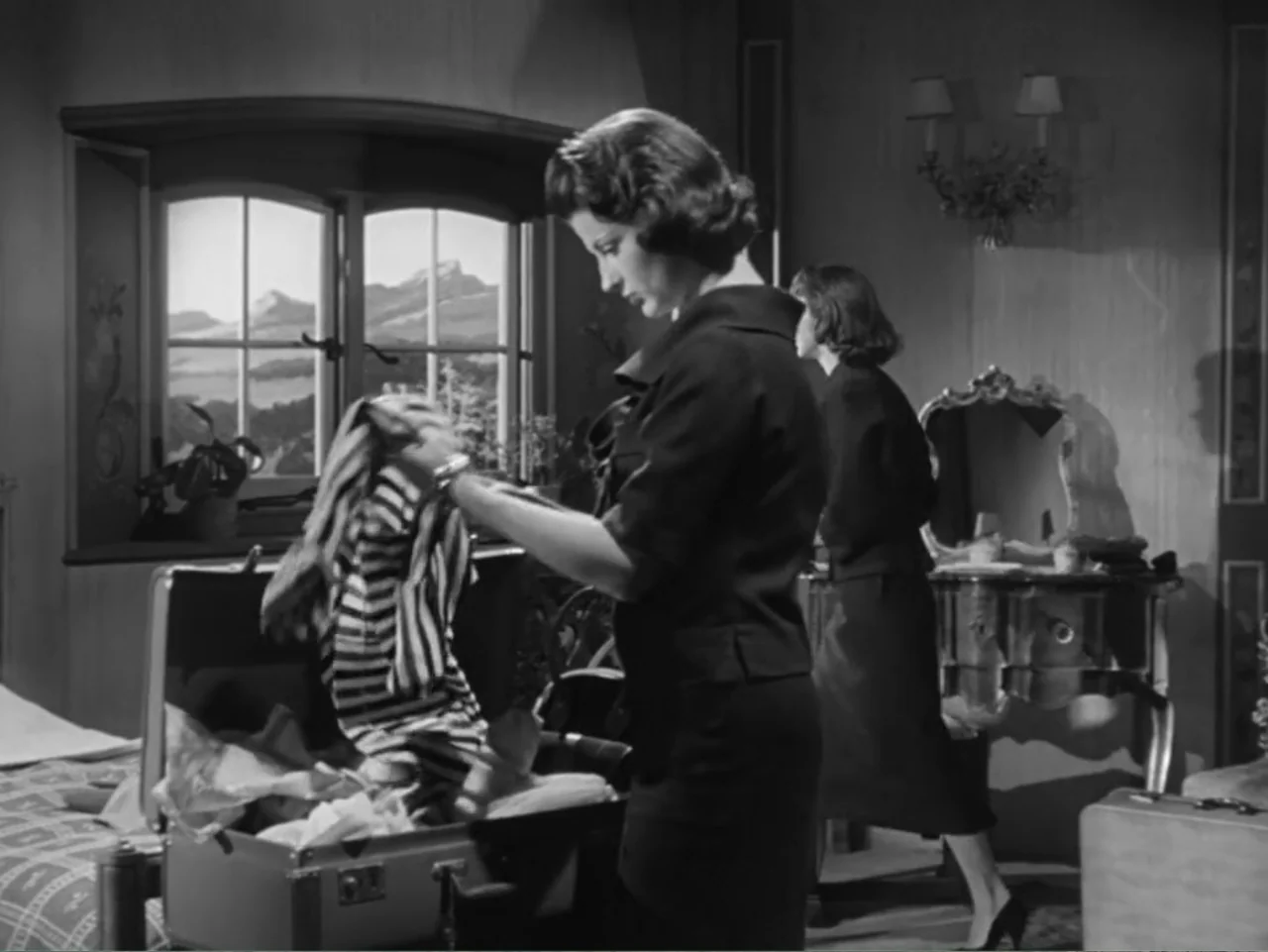 The Crawling Eye (1958) movie still: medium — A mid-century hotel room features a window with a mountain landscape view. A woman packs striped…