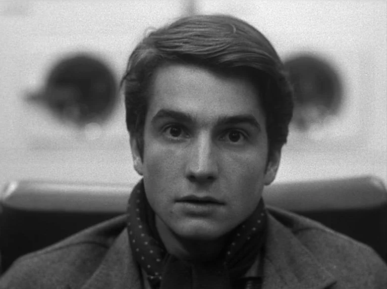 Masculin Féminin (1966) movie still: closeup — A man stands in front of round metal doors within a brightly lit interior. He looks forward with an…