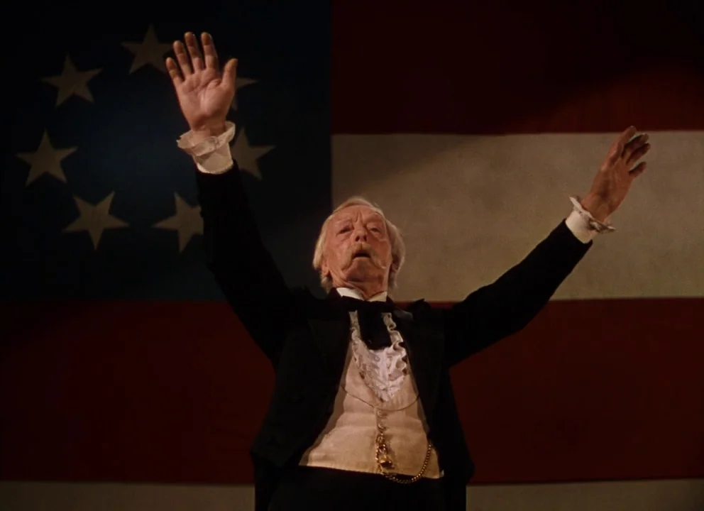 Gone with the Wind (1939) movie still: medium — An elderly man in a suit stands in front of a historical flag. He raises both arms toward the…