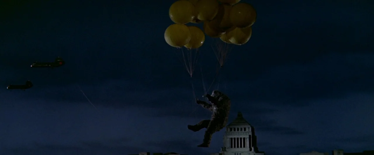 King Kong Vs. Godzilla (1962) movie still: wide — A dark night sky over a cityscape featuring a government building dome. A hairy ape-like figure…