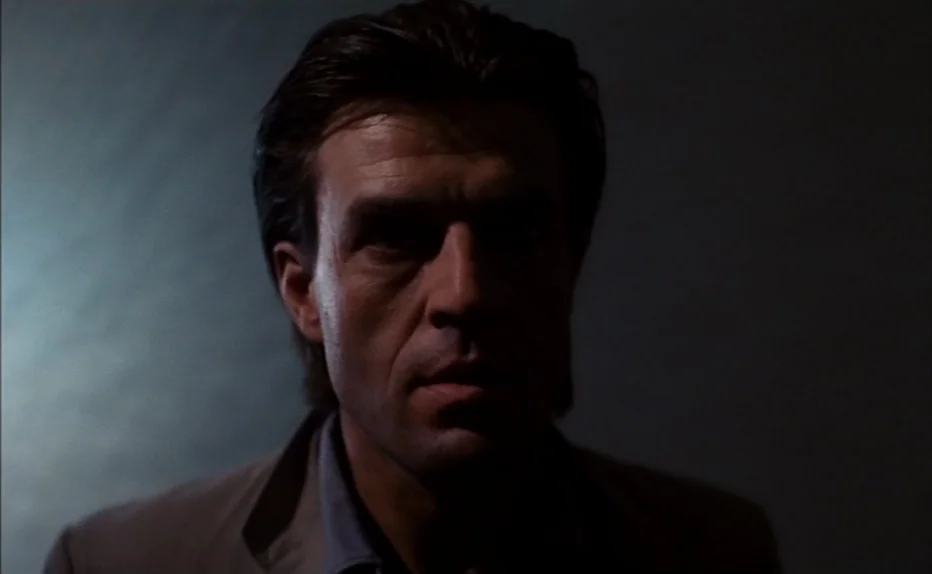The Fourth Man (1983) movie still: closeup — A man stands in a dark room with a neutral gray background. The subject faces the camera directly under…