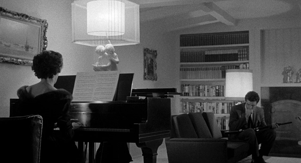 Murder By Contract (1958) movie still: medium — A dark living room features a grand piano, a bookshelf, and a fireplace. A woman plays the piano…