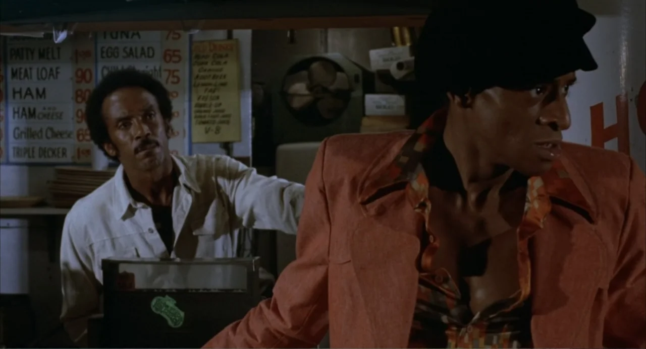 Foxy Brown (1974) movie still: medium — The interior of a vintage diner with a menu board visible in the background. A man in a bright blazer looks…