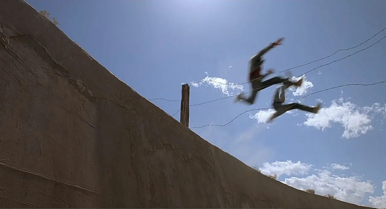 Tremors (1990) movie still: wide — A high, smooth concrete wall runs along a barbed wire fence under a bright sky. Two figures are captured mid-air,…