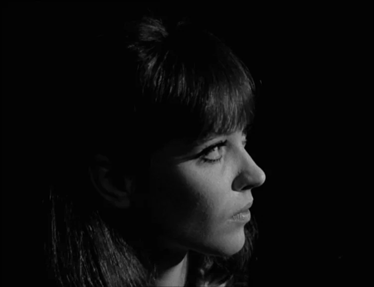 Alphaville (1965) movie still: closeup — A woman stands against a black background with no visible environment details. The lighting emphasizes the…