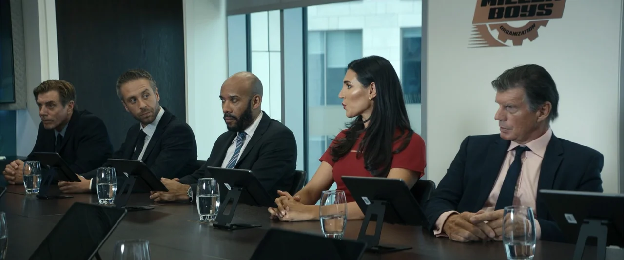 Maintenance Required (2025) movie still: medium — A modern boardroom with a long dark wooden table and large glass windows. Five people in business…