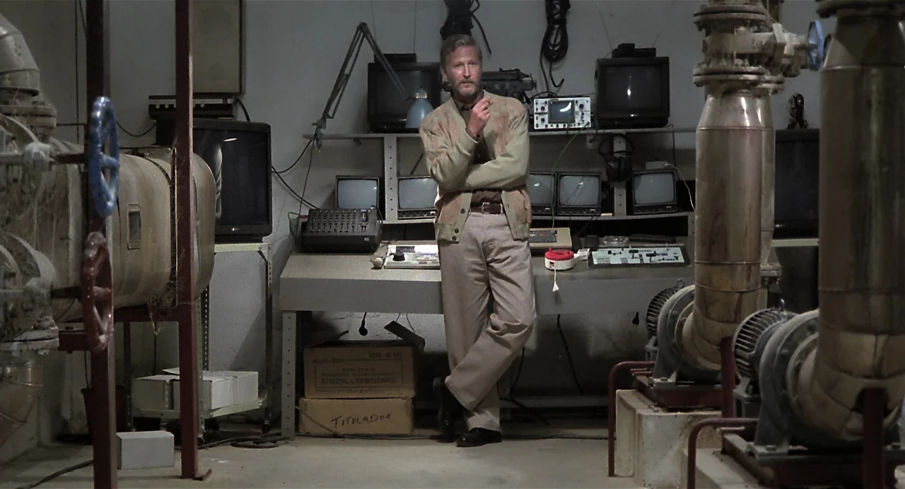 Thesis (1996) movie still: fullbody — A cluttered technical basement features multiple monitors and metal plumbing fixtures arranged symmetrically.…
