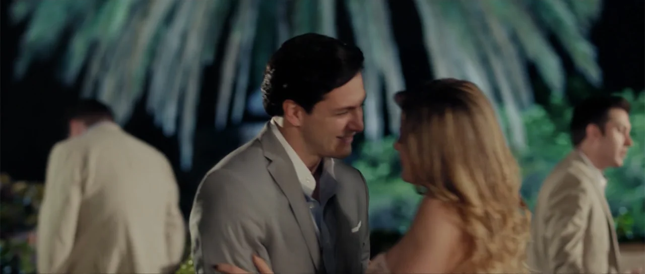 Cindy La Regia (2020) movie still: medium — An outdoor evening scene features a man and woman in formal attire interacting closely. Other figures in…