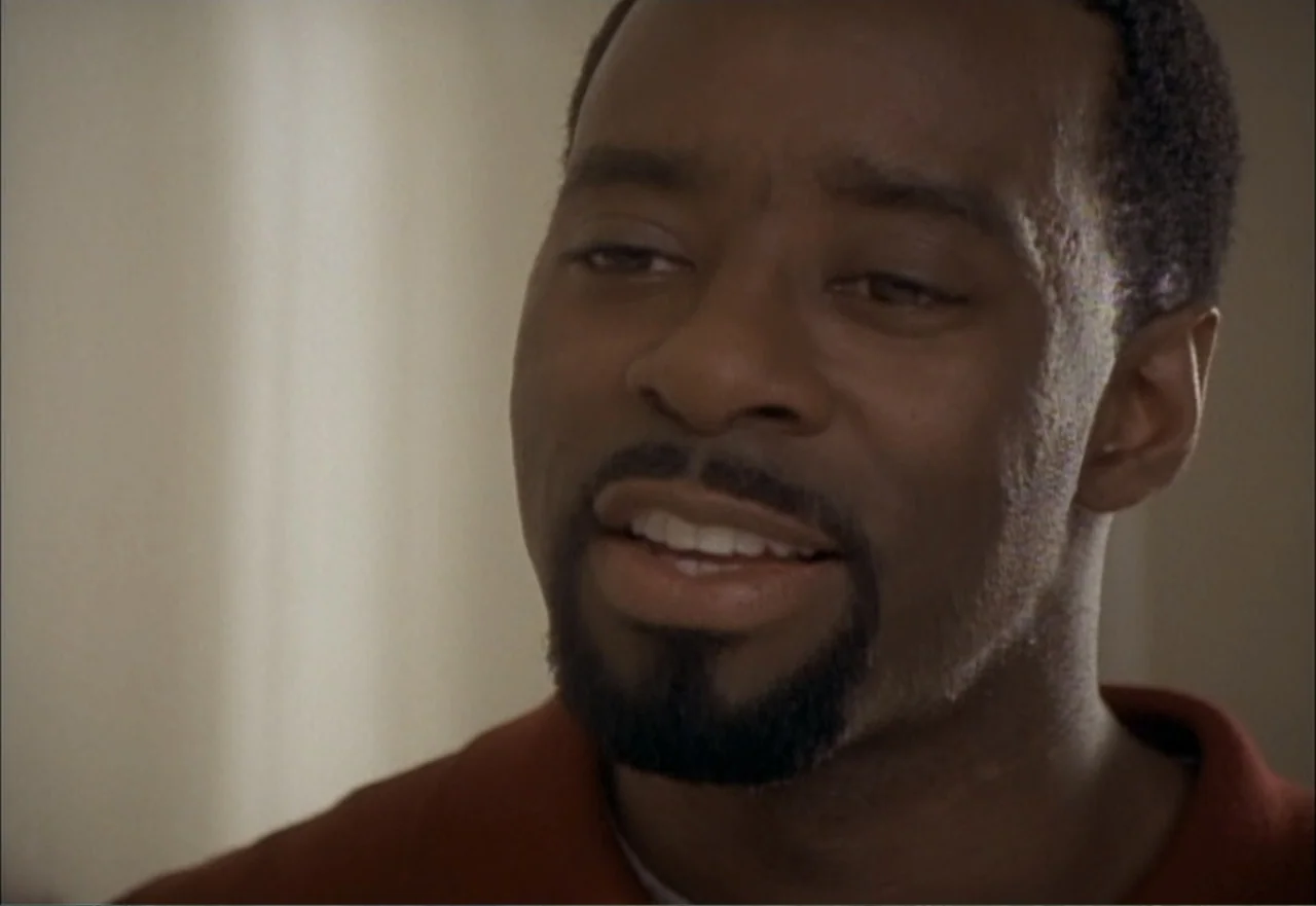 Ambushed (1998) movie still: closeup — A man stands indoors against a blurred neutral background. He looks slightly away from the lens while smiling…