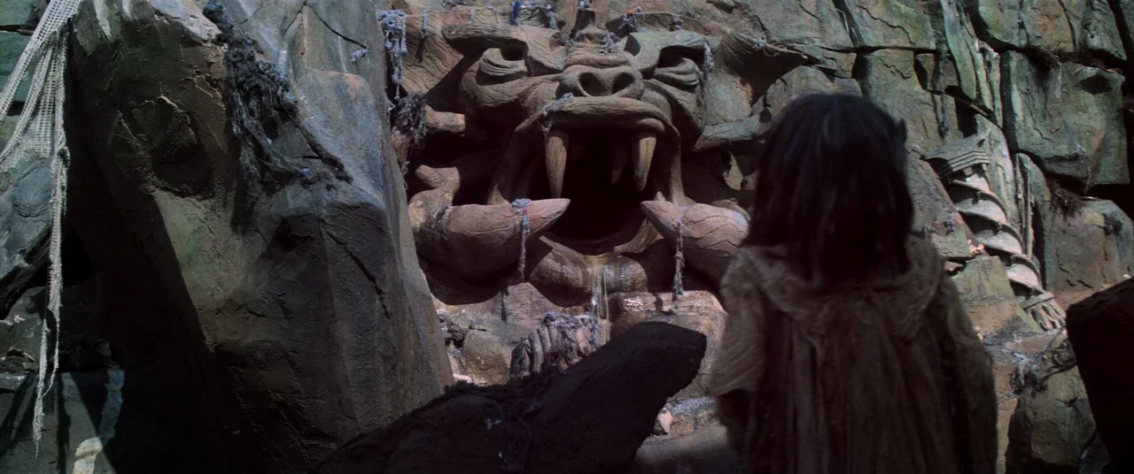 The Dark Crystal (1982) movie still: medium — A dark rocky grotto features a large stone relief sculpture of a creature's face. A small figure…
