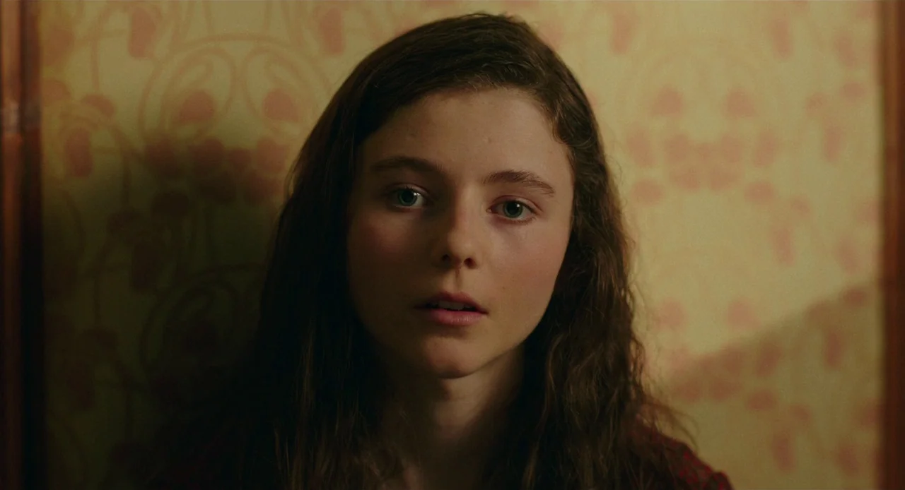 Jojo Rabbit (2019) movie still: closeup — A close-up shot features a girl positioned against a light-colored wallpaper with repeating patterns. She…