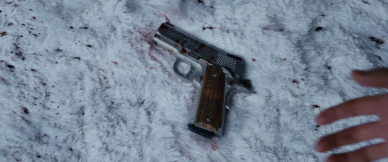 Under The Silver Lake (2018) movie still: closeup — A high-angle close-up shows a silver semi-automatic pistol resting on a white shag rug. The…
