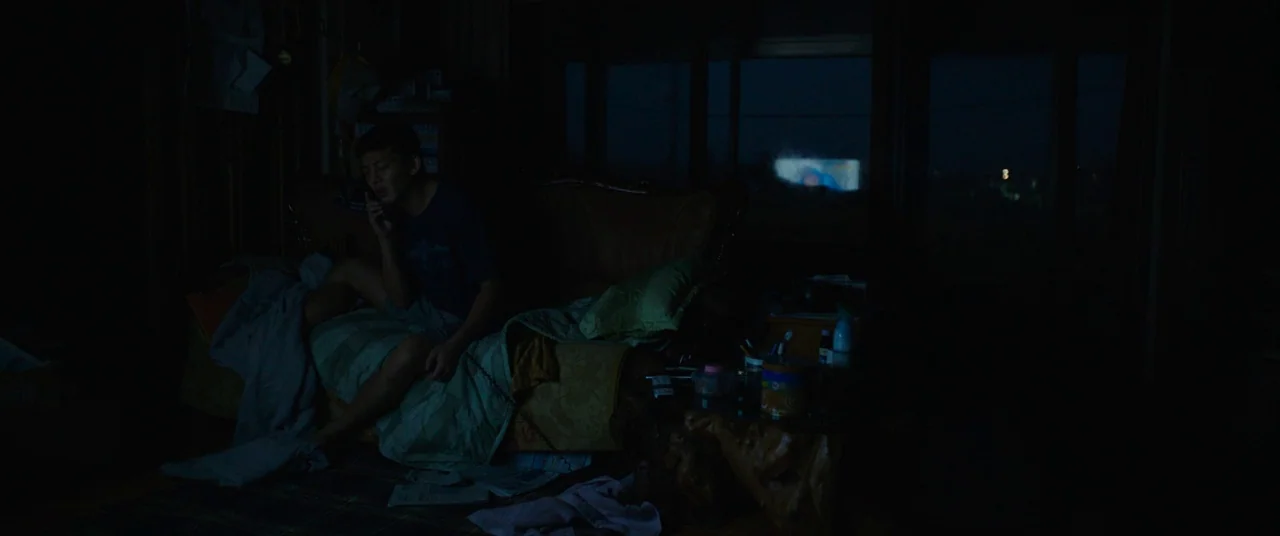 Burning (2018) movie still: medium — A dimly lit room at night with a man sitting on a sofa. He is holding a mobile phone to his ear in the dark…