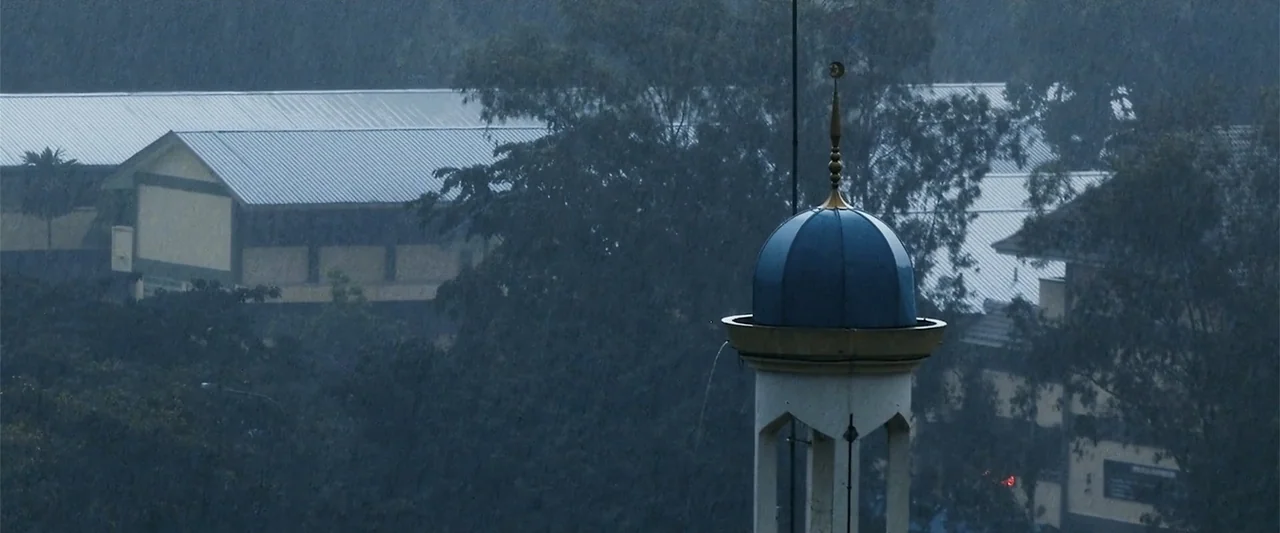 The Kleptocrats (2018) movie still: establishing — A mosque minaret top stands prominently against a backdrop of trees and building roofs. Heavy…