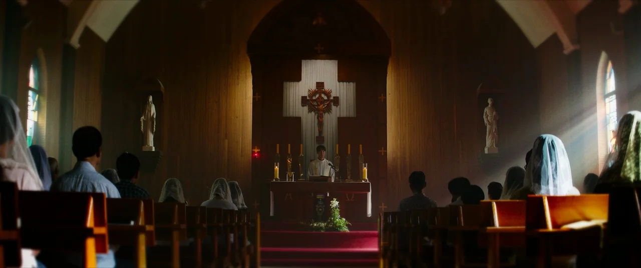 The Divine Fury (2019) movie still: wide — A wooden church interior with a central altar features a large wooden crucifix above. A congregation sits…