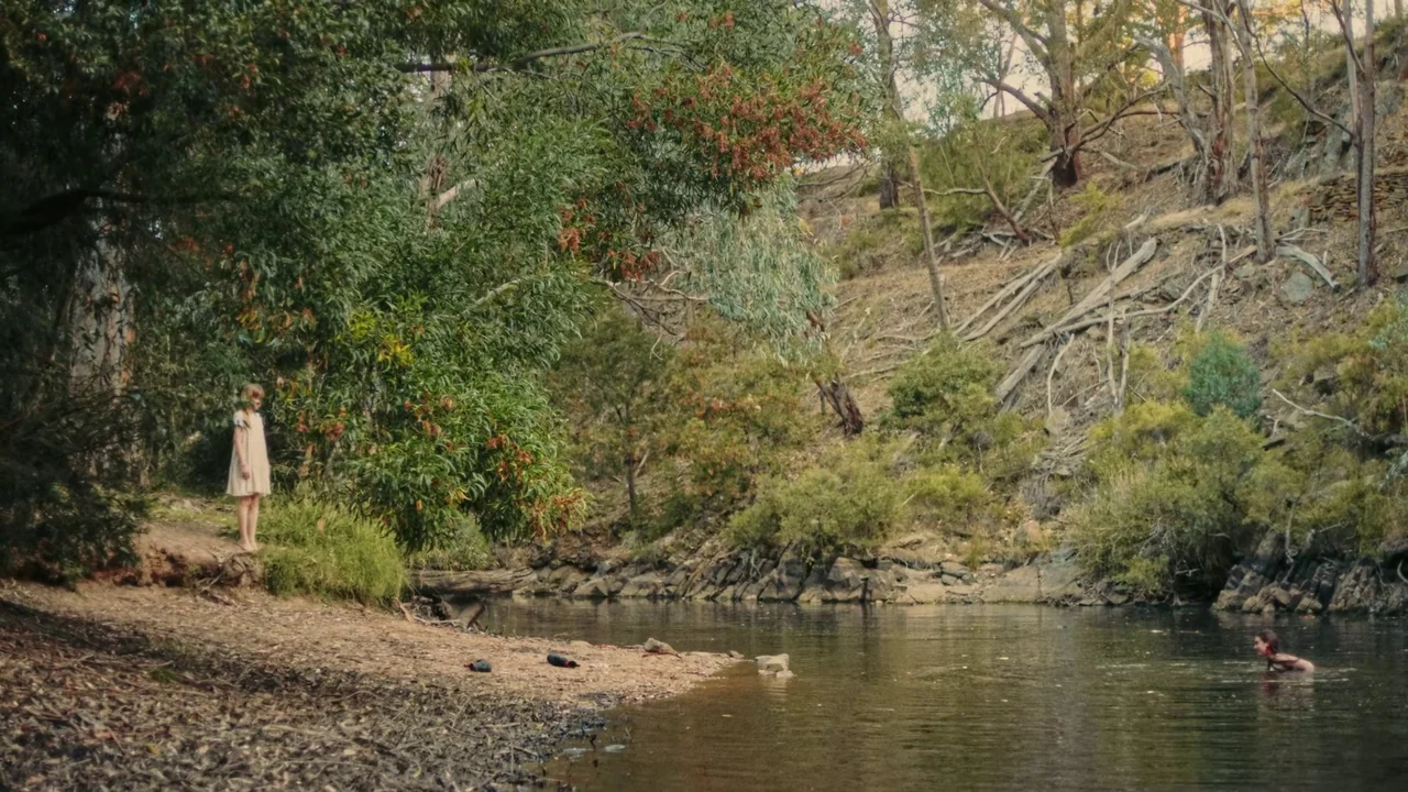 My First Summer (2020) movie still: wide — A rural creek bank is surrounded by eucalyptus trees and a dry hillside. One girl stands on the sandy…