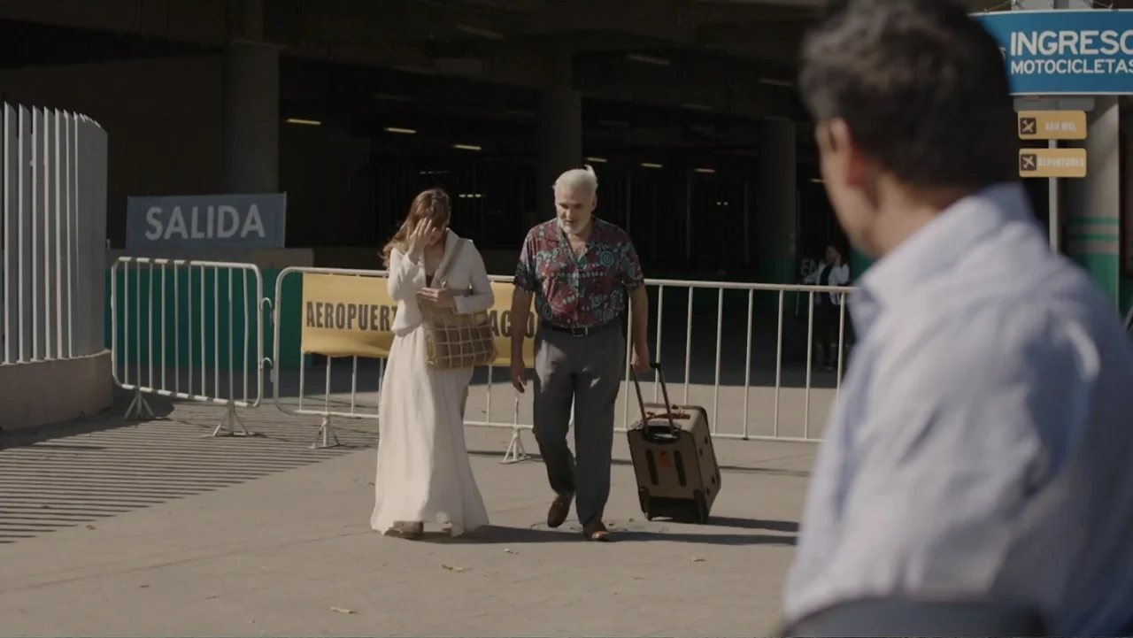 Por Amor al Money (2023) movie still: fullbody — An outdoor airport pickup zone features metal crowd-control barricades and informational signs. A…