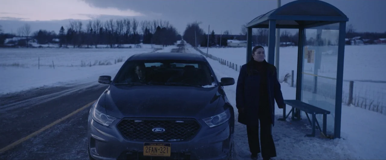 The Lie (2018) movie still: wide — A snowy rural landscape features a dark sedan stopped beside a roadside bus shelter. A woman in a dark coat walks…