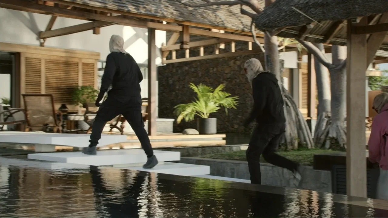 Unknown #1 movie still: fullbody — A modern outdoor living space features wooden beams and stone stepping stones over water. Two figures in black…