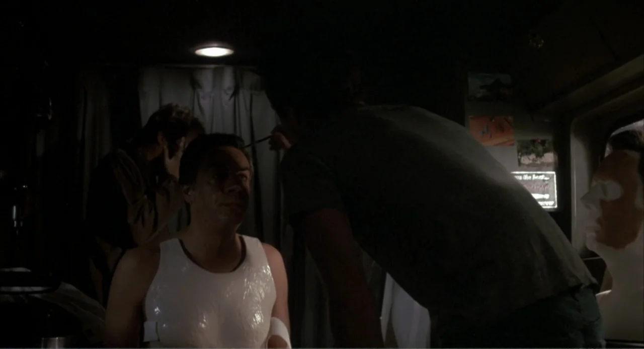 F/X (1986) movie still: medium — A dimly lit interior space with fabric curtains and assorted items in the background. A man sits wearing a white…