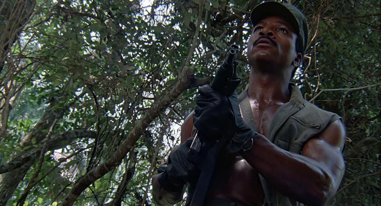 Predator (1987) movie still: medium — A dense, overgrown jungle with tangled branches and thick foliage filling the frame. A muscular soldier in…