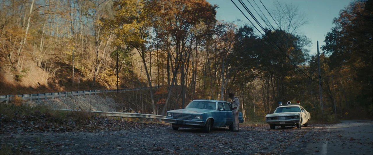 I'm Your Woman (2020) movie still: wide — A rural paved road winds through a dense forest during the autumn season. A state trooper stands beside a…