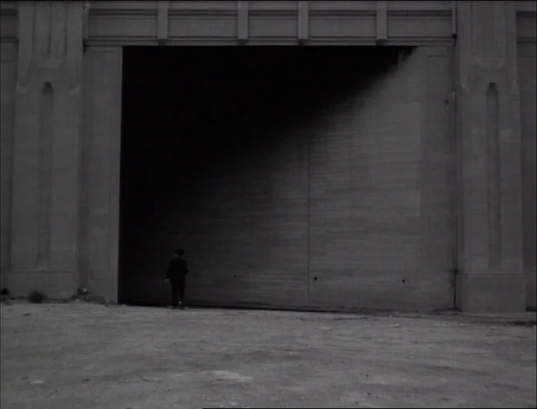 Eraserhead (1977) movie still: wide — A monolithic concrete structure with a massive, dark, shadowed portal dominates the frame. A single small…
