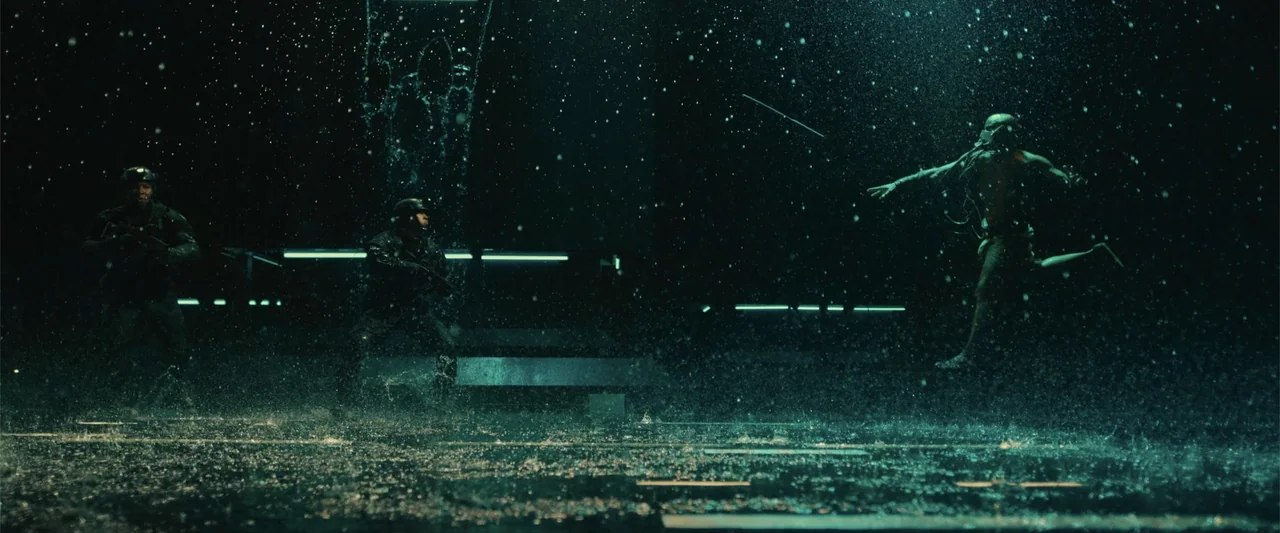 Black Adam (2022) movie still: wide — A dark, rainy outdoor area at night illuminated by distant blue neon lights. Two soldiers in tactical armor…