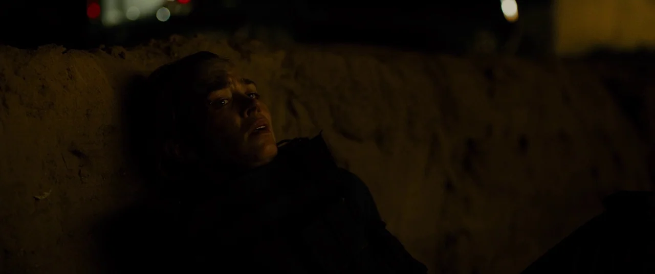 Sicario (2015) movie still: closeup — A person leans against a dark, rough-textured earthen mound in total darkness. The subject is partially…