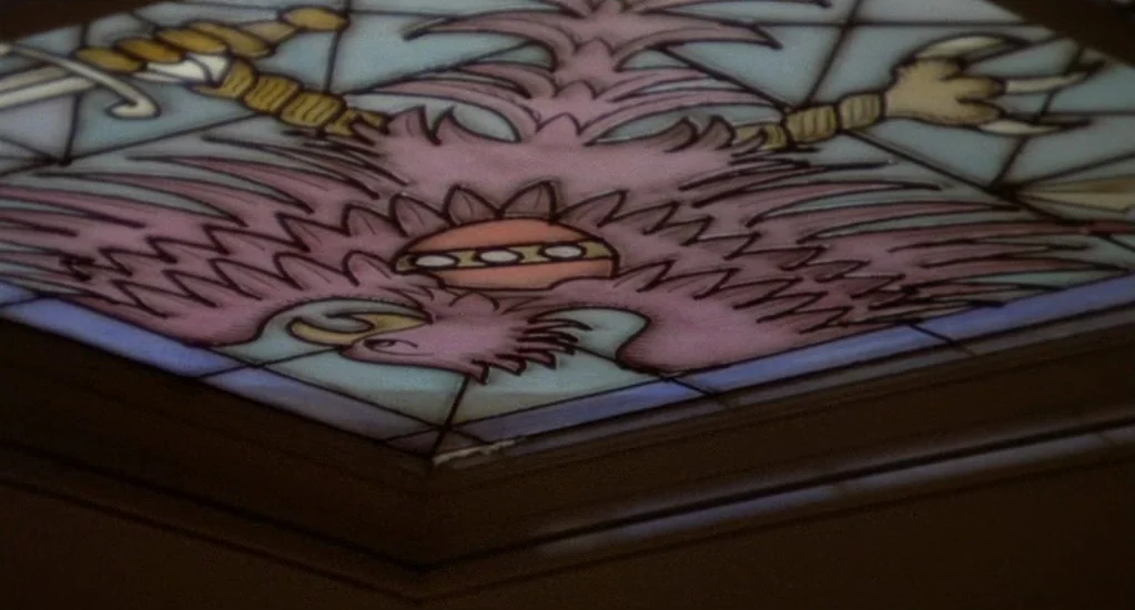 Demons of the Mind (1972) movie still: closeup — A close-up view of a decorative stained glass panel depicting a heraldic eagle design. The glass is…