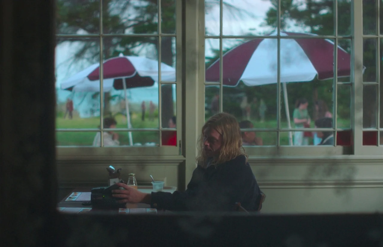 Nitram (2021) movie still: medium — A diner interior overlooks an outdoor patio space with large umbrellas through grid windows. A lone man with…