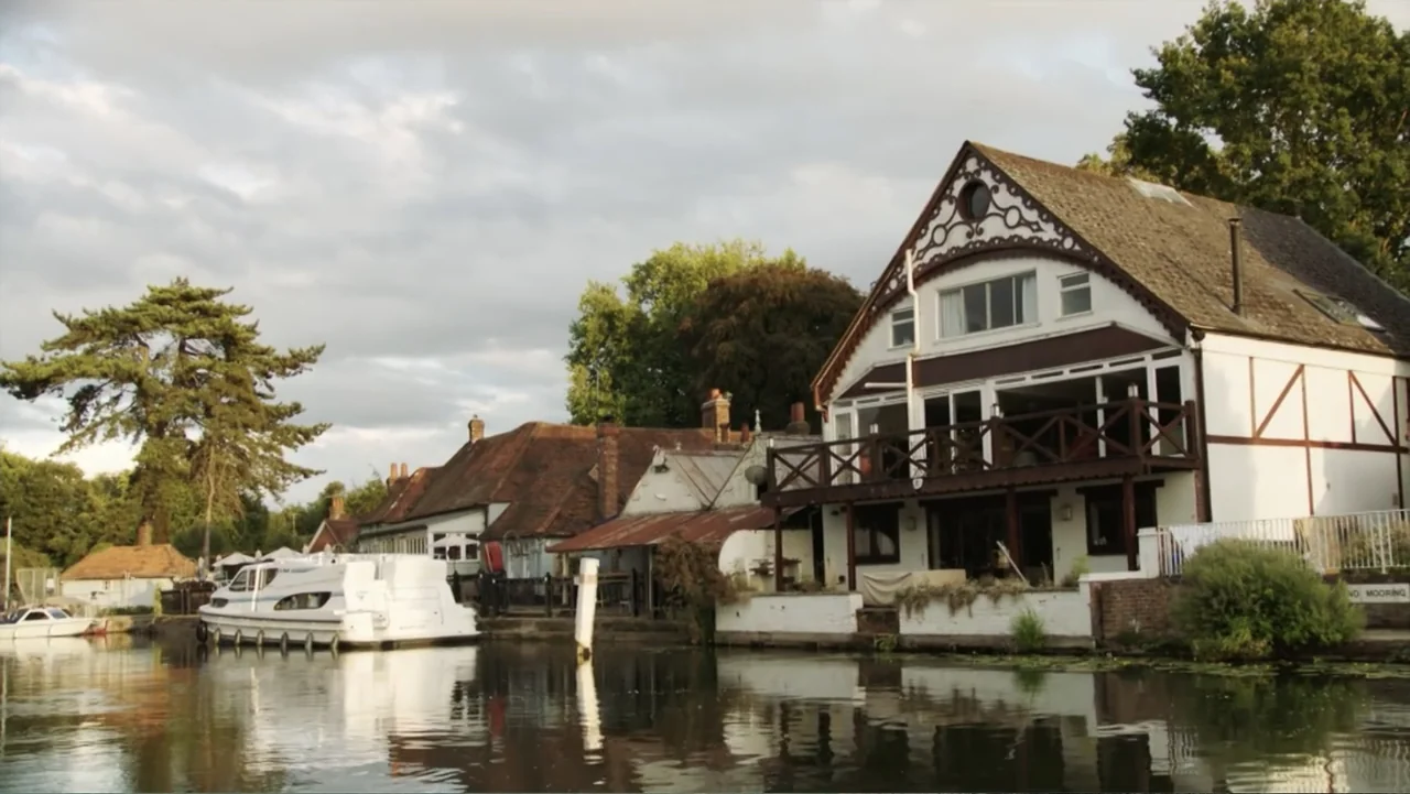Becoming Led Zeppelin (2025) movie still: establishing — A row of historic white buildings with timber frames sits along a calm river. A white…