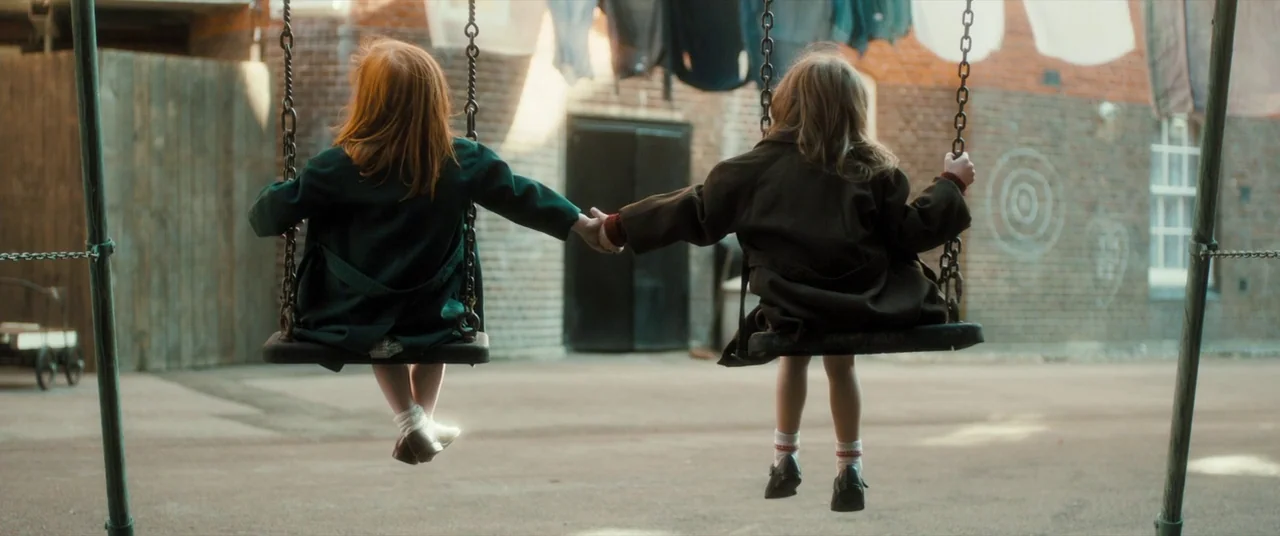 Ginger & Rosa (2012) movie still: fullbody — A paved area behind a brick building features laundry hanging between walls. Two young children face…