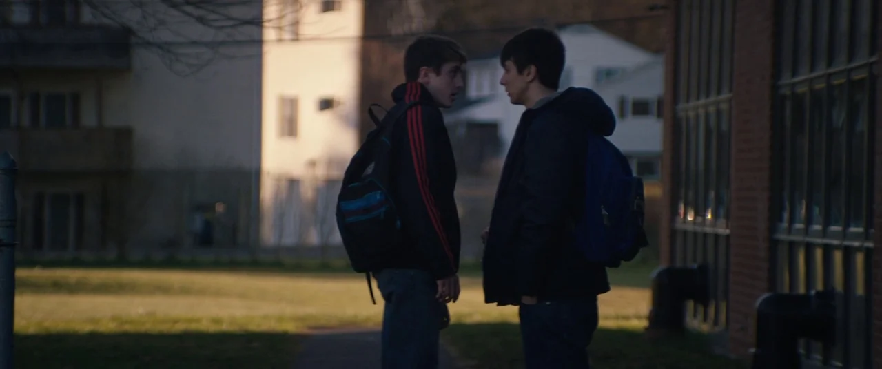Super Dark Times (2017) movie still: fullbody — Two teenage boys stand talking on a lawn during the late afternoon. A large brick building and…