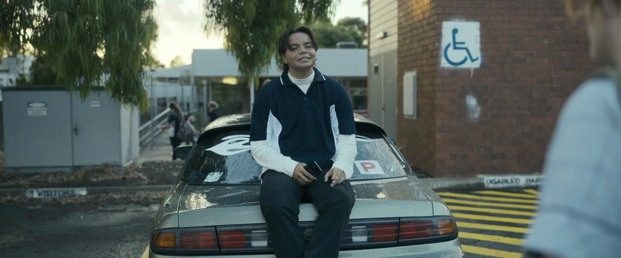 Talk to Me (2023) movie still: medium — A person sits on the trunk of a silver sedan in a paved parking lot. The person is holding a smartphone and…