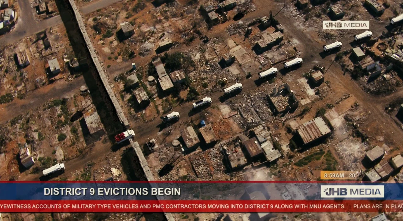 District 9 (2009) movie still: establishing — An aerial view shows a sprawling slum with dilapidated structures and unpaved roads. A line of white…