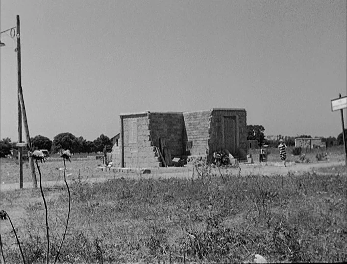 Nights of Cabiria (1957) movie still: wide — A desolate open field features a small, boxy, unadorned stone building in the center. A lone person in…