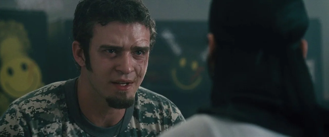 Southland Tales (2006) movie still: medium — A dimly lit room with walls decorated by yellow smiley face signs. A man wearing camouflage gear stares…