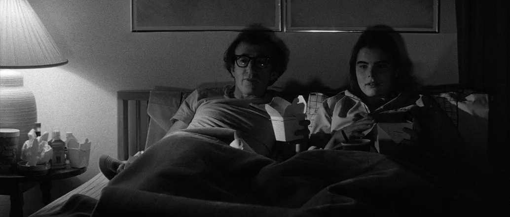 Manhattan (1979) movie still: medium — A dark bedroom with two people lying in bed under a blanket. The man and woman hold white paper takeout…