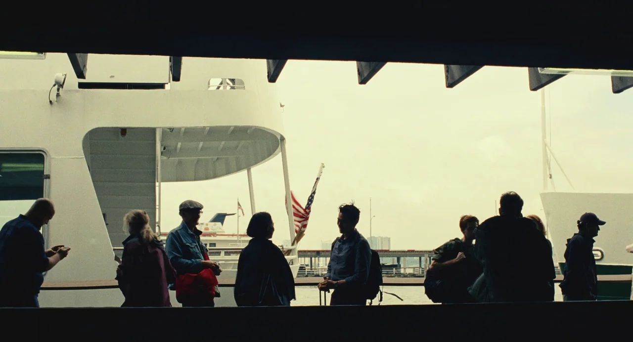 Past Lives (2023) movie still: wide — A group of passengers stands on the deck of a large white ferry vessel. They face toward the water under a…