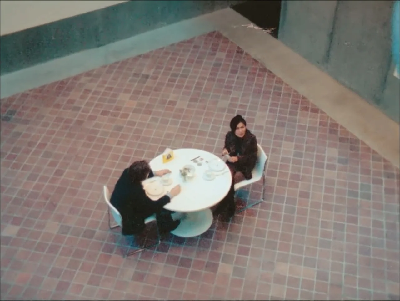 One to One: John & Yoko (2025) movie still: wide — A minimalist gallery space features a small round table set on a tiled floor. A man and a woman…